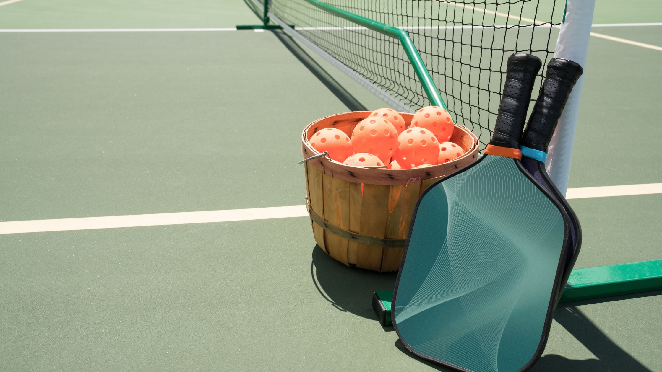 two paddles and a basket of pickleballs on a court