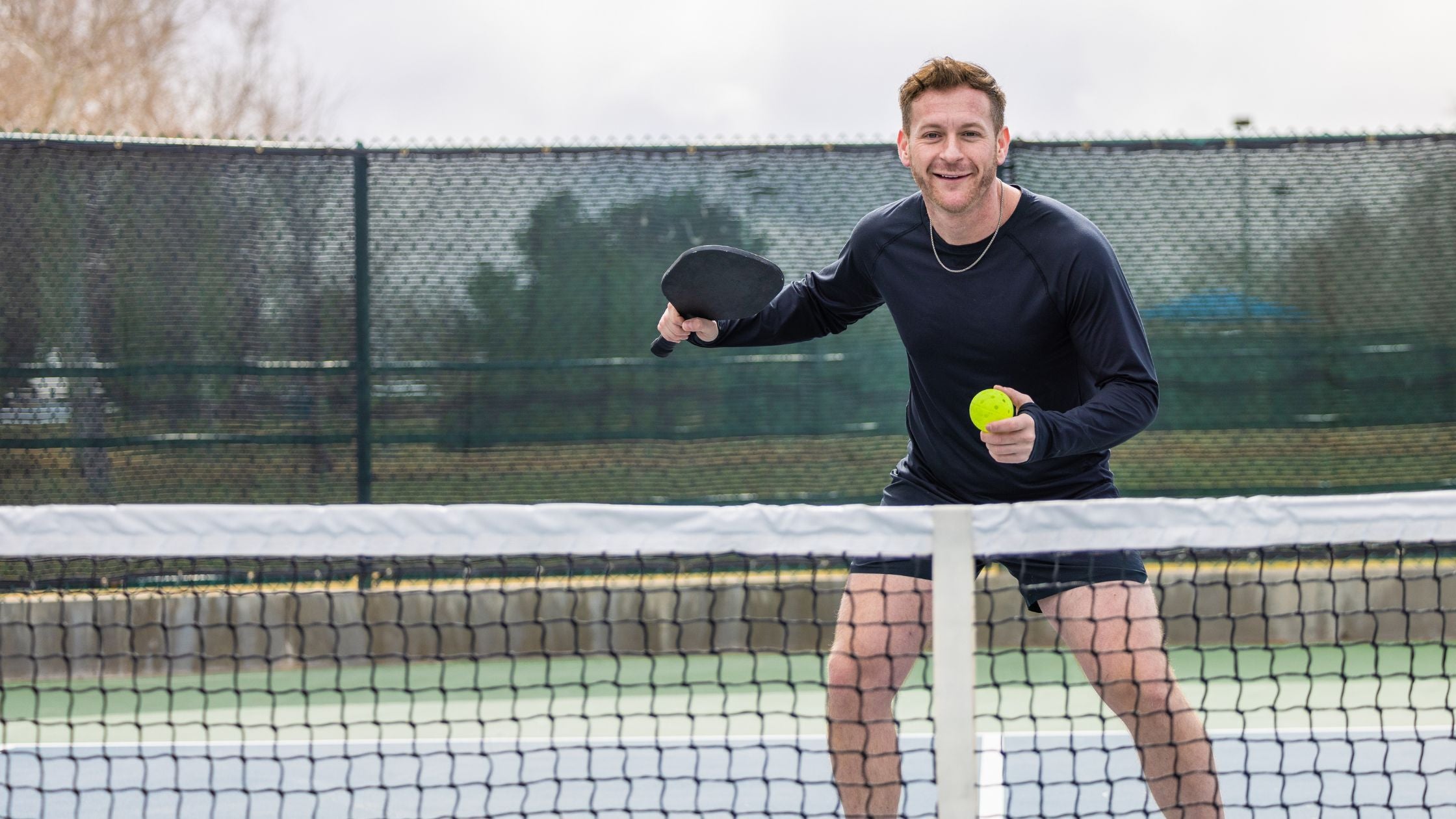 a man smiling while holding a pickleball paddle and ball